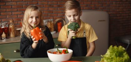 Cute girl and boy with fresh pepper and broccoli in hands standing near desk with assorted vegetables and salad bowl during vegetarian meal preparation