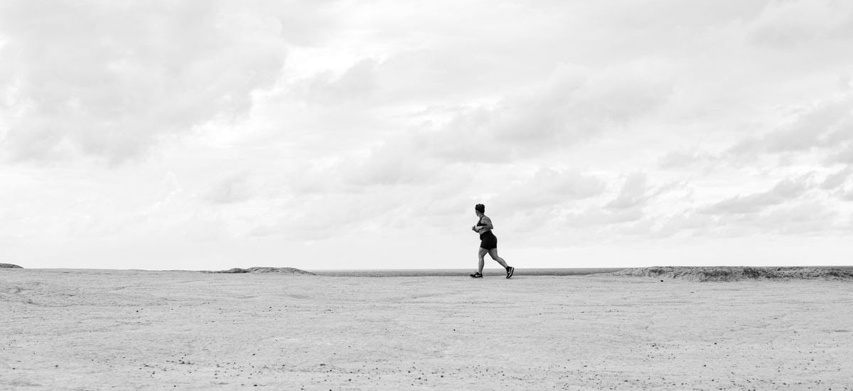 A lone runner traverses a vast, empty Brazilian beach under dramatic cloudy skies in black and white.